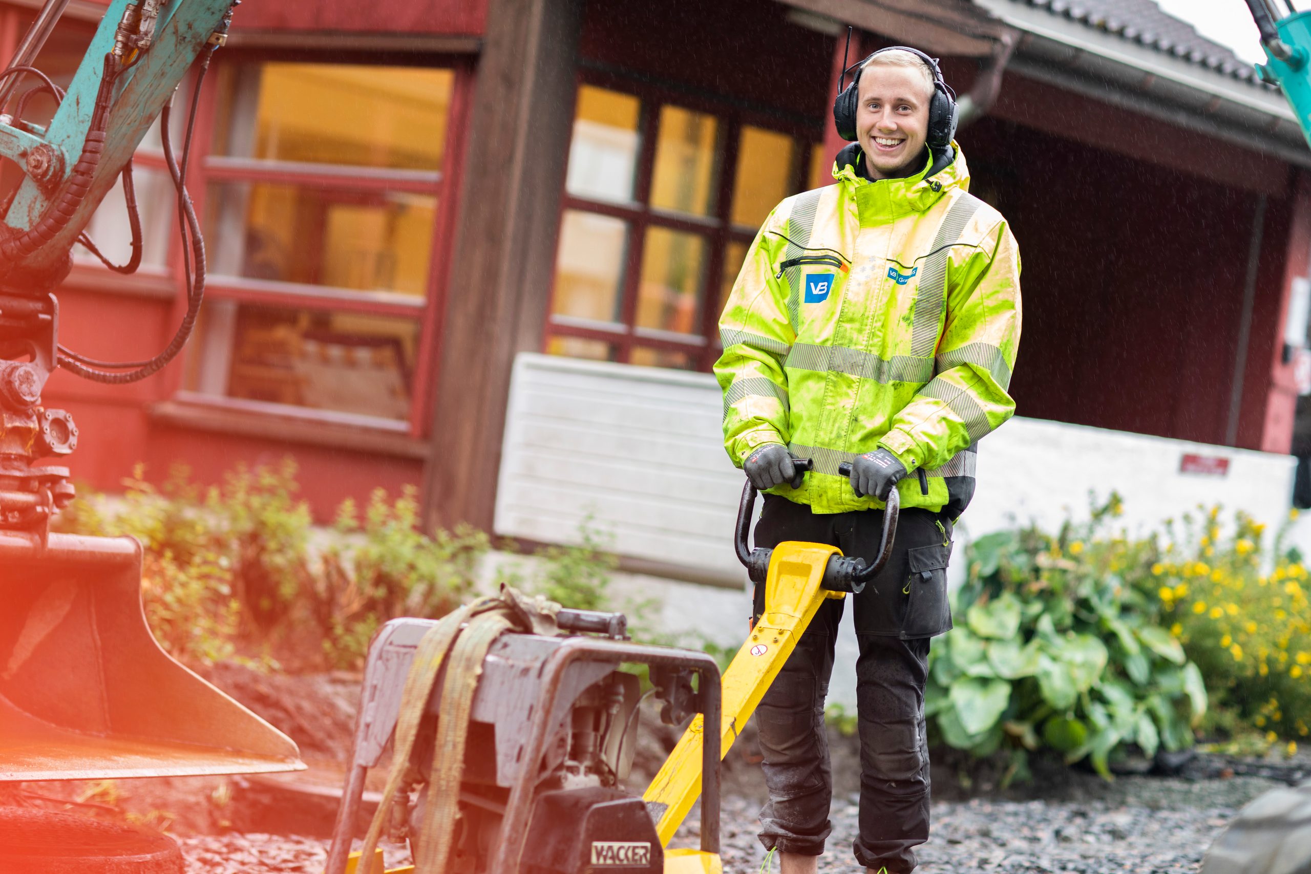 Anleggsrørlegger med refleksjakke og hørselsvern jobber med platekompaktor/hoppetusse ved en gravemaskin utenfor bolig.
