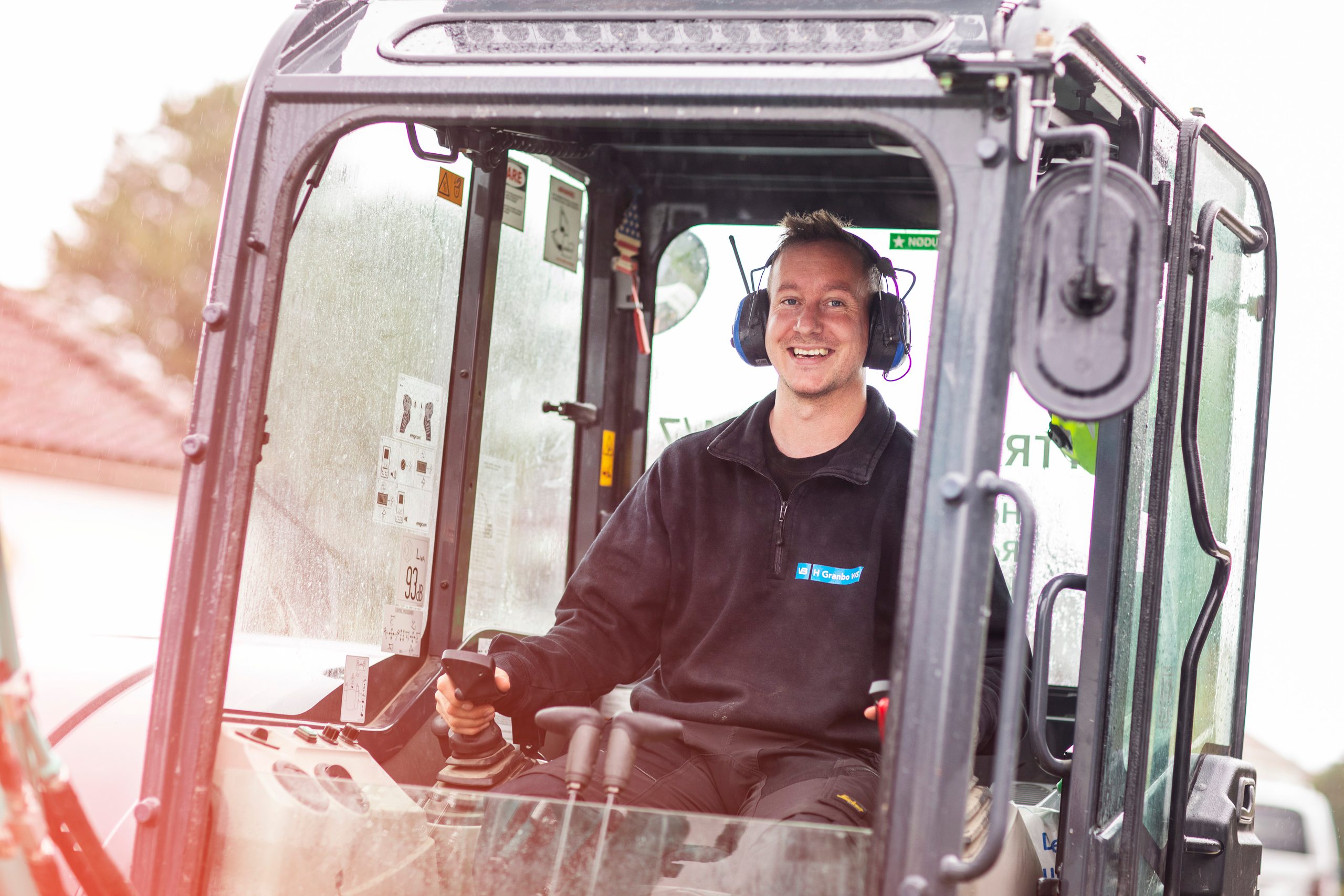 Anleggsrørlegger med arbeidsjakke, stort smil og hørselsvern med radio i gravemaskin.